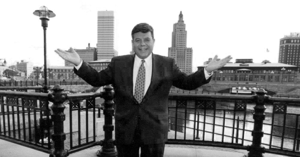 A black and white photo of Cianci standing in front of the Providence River lagoon with the city's skyline in the background. Cianci was instramental in the creation of the lagoon as part of the 'Providence Renaissance' which he is credited with engineering.